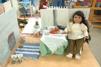 Doll sitting on chair in kitchen with rag rug and tableclothes.